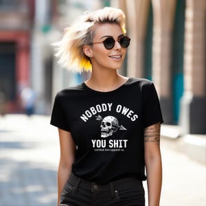 May include: Black t-shirt with the slogan "NOBODY OWES YOU SHIT" in white, featuring a skull pierced by an arrow. The shirt also includes "combat iron apparel co." at the bottom. The model is wearing sunglasses.