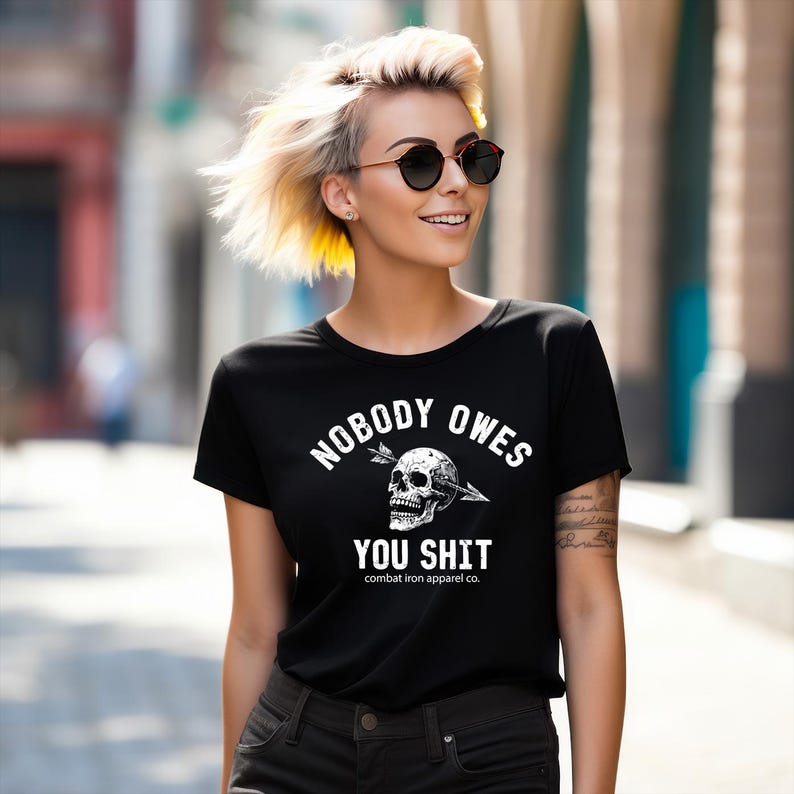 May include: Black t-shirt with the slogan "NOBODY OWES YOU SHIT" in white, featuring a skull pierced by an arrow. The shirt also includes "combat iron apparel co." at the bottom. The model is wearing sunglasses.
