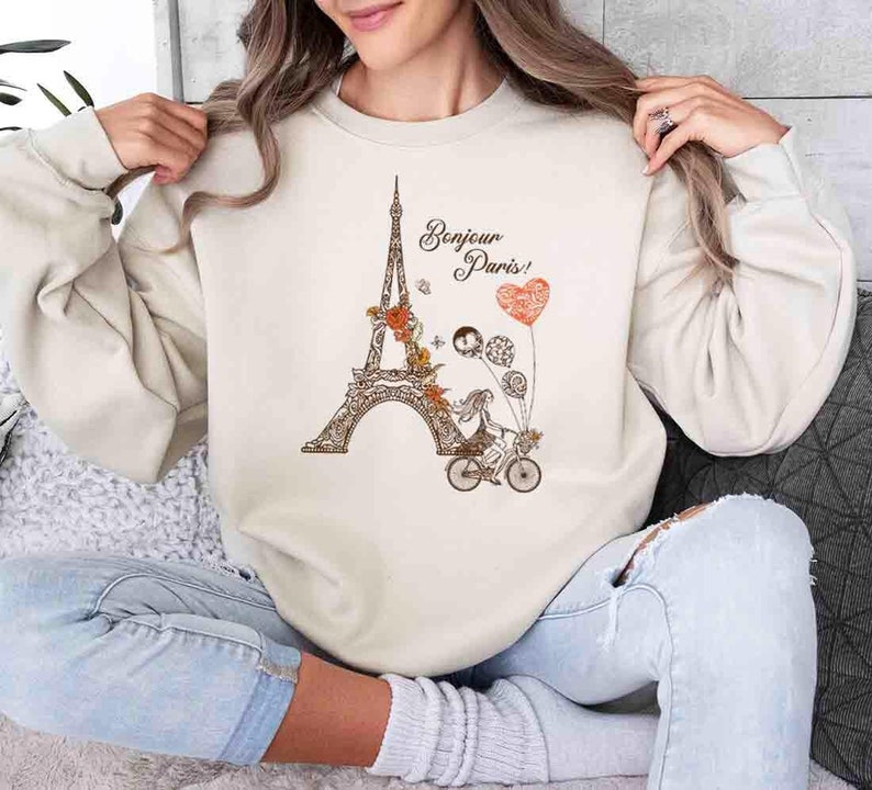 Pode incluir: Uma camisa de moletom bege com um gr&aacute;fico da Torre Eiffel, uma mulher andando de bicicleta e o texto "Bonjour Paris!" em letras marrons.