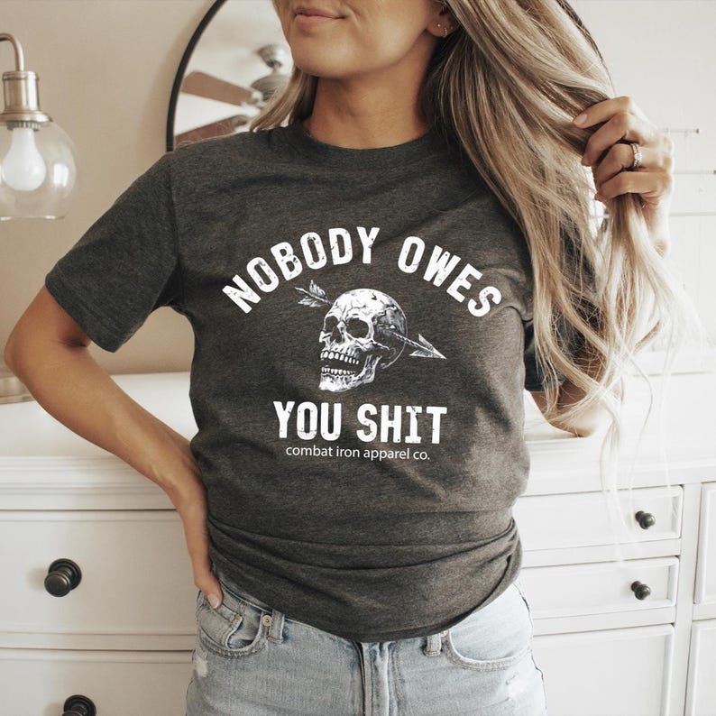 May include: Dark grey t-shirt with the slogan "NOBODY OWES YOU SHIT" in white, featuring a skull pierced by an arrow. The shirt also includes the text "combat iron apparel co."