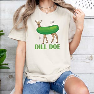 Puede incluir: Una camiseta beige claro con una imagen de dibujos animados de un ciervo que lleva un disfraz de pepinillo verde. El texto "DILL DOE" est&aacute; impreso debajo de la imagen.