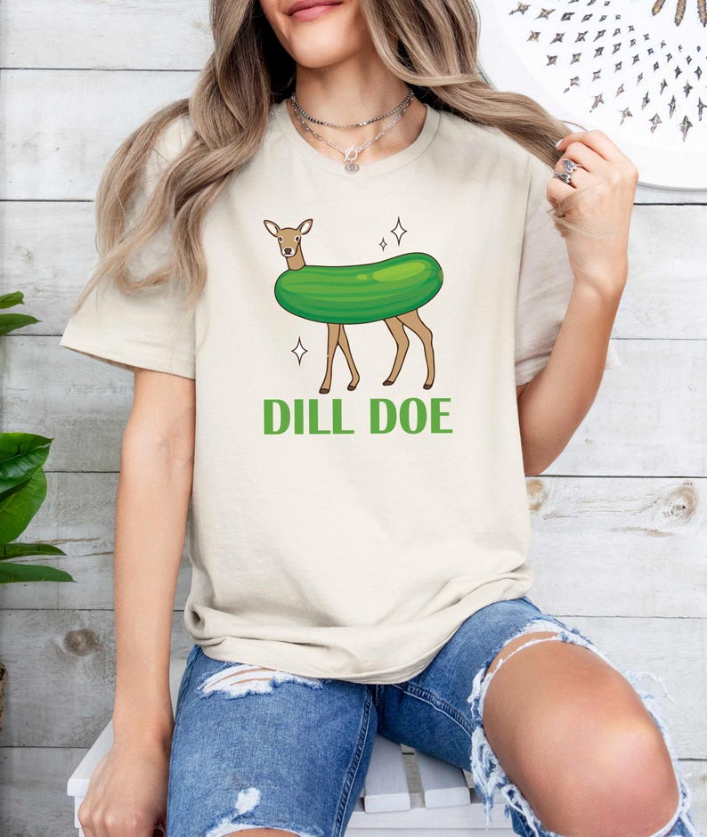 Puede incluir: Una camiseta beige claro con una imagen de dibujos animados de un ciervo que lleva un disfraz de pepinillo verde. El texto "DILL DOE" est&aacute; impreso debajo de la imagen.