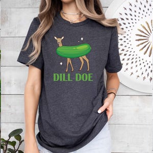 Puede incluir: Una camiseta gris oscuro jaspeada con un gr&aacute;fico de un ciervo que lleva un disfraz de pepinillo. El texto "Dill Doe" est&aacute; impreso debajo de la imagen.