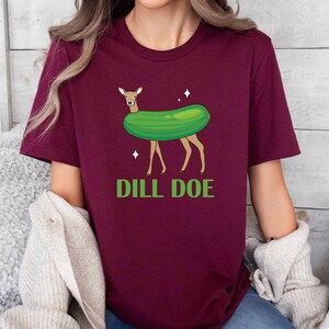 Puede incluir: Una camiseta de color granate con un gr&aacute;fico de un ciervo que lleva un pepinillo verde y el texto "DILL DOE" en verde.
