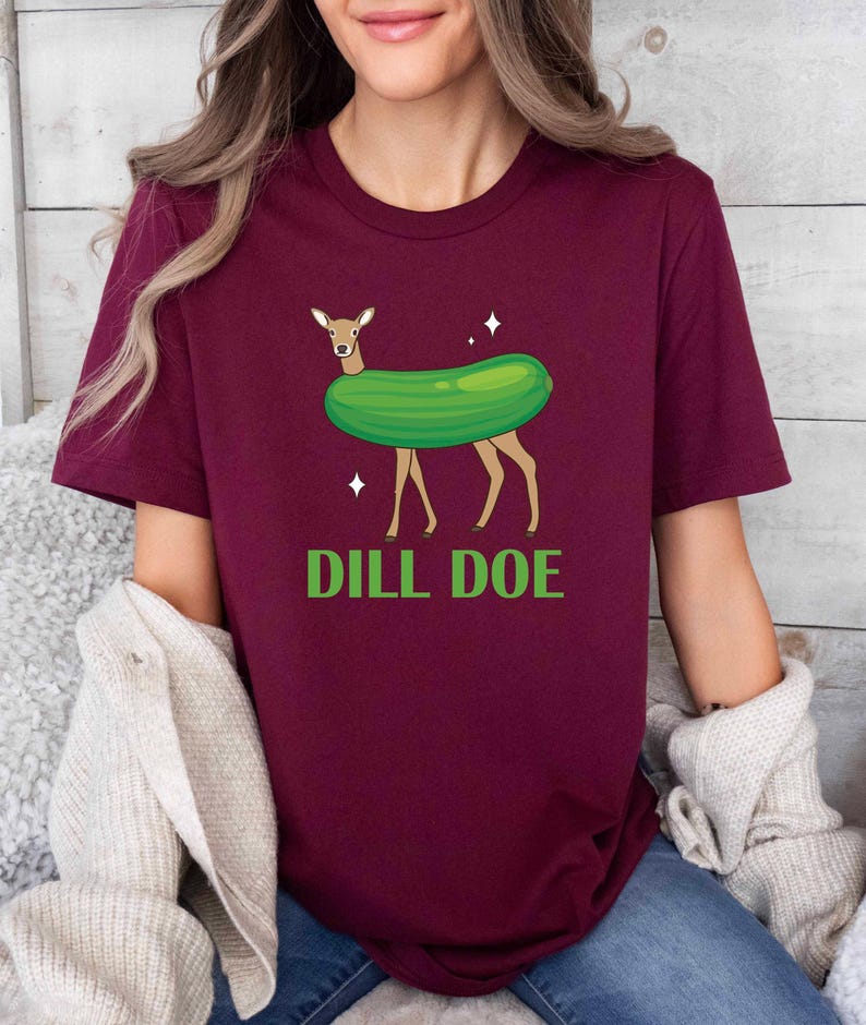 Puede incluir: Una camiseta de color granate con un gr&aacute;fico de un ciervo que lleva un pepinillo verde y el texto "DILL DOE" en verde.
