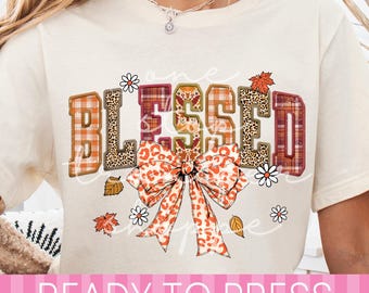 May include: A cream-colored short-sleeve t-shirt featuring the word BLESSED in large block letters with plaid and leopard print patterns. A large orange leopard print bow sits below the text, surrounded by small white daisies and autumn leaves.