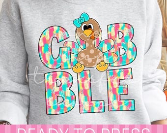 May include: A light gray sweatshirt featuring a graphic print of a brown turkey with a teal bow. The text GIBBLE is displayed in large letters with a colorful pattern of teal, pink, and yellow brushstrokes and black dots on a white background.