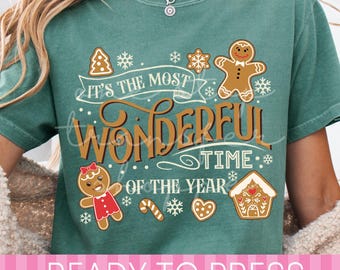 May include: A green short-sleeve t-shirt featuring a graphic print with the text "IT'S THE MOST WONDERFUL TIME OF THE YEAR" in brown and white. The design includes gingerbread people, a gingerbread house, snowflakes, and candy canes.