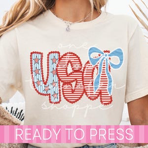May include: Cream-colored t-shirt with a patriotic "USA" design in red, white, and blue. The design includes stars, stripes, and a blue bow. The words "one" and "shop" are written in cursive. The text "READY TO PRESS" is at the bottom.
