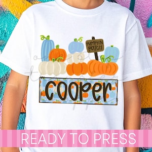 May include: A white short-sleeved t-shirt featuring a graphic of stacked pumpkins in shades of orange, white, and light blue. A brown sign reads PUMPKIN PATCH. Below, a rectangular box with a blue pumpkin pattern contains the name COOPER.