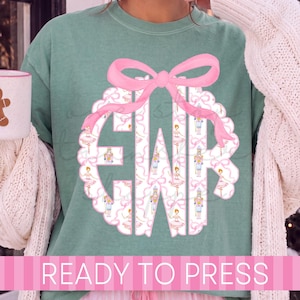 May include: A sage green t-shirt featuring a large, scalloped-edge monogram with the letters GWR. The monogram is filled with a pattern of pink bows and nutcracker figures, topped with a large pink bow. The text READY TO PRESS is at the bottom.
