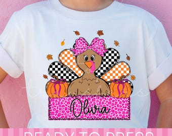 May include: A white t-shirt featuring a graphic of a brown turkey wearing a pink leopard print bow. The turkey is surrounded by orange and black checkered feathers, two orange pumpkins, and the name Olivia in a pink leopard print rectangle.