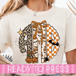May include: A cream-colored t-shirt featuring a large, circular monogram design with the letters AMB. The A has a leopard print, the M features a pumpkin and bat pattern with a brown bow, and the B has an orange and white checkerboard pattern.