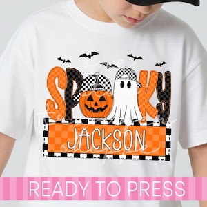 May include: A white t-shirt featuring a graphic print with the word SPOOKY in orange and black checkered letters. The design includes a jack-o'-lantern and a ghost wearing checkered caps, black bats, and an orange checkered box with the name JACKSON.