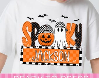 May include: A white t-shirt featuring a graphic print with the word SPOOKY in orange and black checkered letters. The design includes a jack-o'-lantern and a ghost wearing checkered caps, black bats, and an orange checkered box with the name JACKSON.