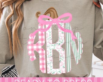 May include: A grey long-sleeve sweatshirt featuring a large graphic of a pumpkin with a pink bow. The pumpkin is filled with a monogram design in pink gingham, a white pumpkin pattern, and green stripes. The text READY TO PRESS is at the bottom.