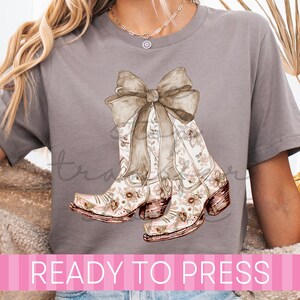 May include: A gray t-shirt featuring a watercolor design of cowboy boots with floral patterns and a large bow. The boots have a brown heel and are decorated with pink and brown flowers. The text "READY TO PRESS" is displayed in pink.