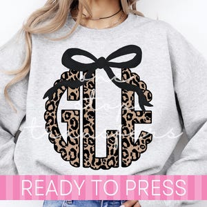 May include: A gray sweatshirt featuring a leopard print monogram with a black bow. The monogram is surrounded by a wreath design. The text "READY TO PRESS" is displayed on a pink banner at the bottom of the image.