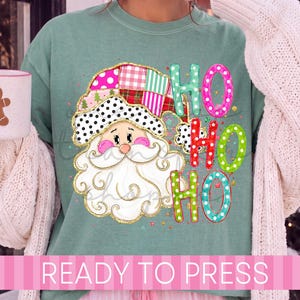 May include: A sage green t-shirt featuring a graphic of Santa Claus wearing a colorful patchwork hat with polka dots and plaid patterns. The text HO HO HO is printed in bright, dotted letters next to the Santa illustration on the front.
