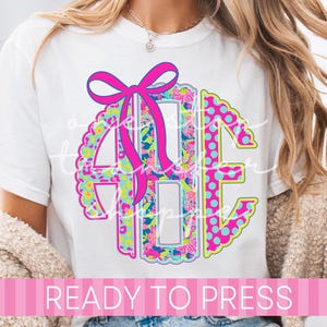 May include: White t-shirt featuring a colorful monogram design with a pink bow. The monogram includes floral and polka dot patterns in shades of blue, pink, and yellow. The text "READY TO PRESS" is displayed on a pink banner.