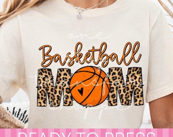 Puede incluir: Una camiseta de color crema con el texto "Basketball MOM" en un diseño gráfico. La palabra "Basketball" está en letra cursiva naranja, mientras que "MOM" está en letras de bloque con estampado de leopardo. Un balón de baloncesto sustituye a la "O".
