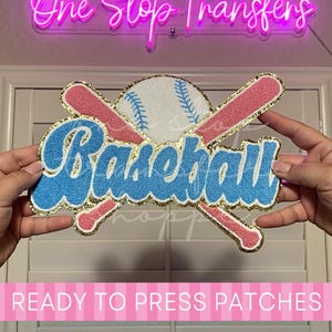 May include: A chenille patch featuring the word Baseball in blue with a white outline, layered over two crossed pink baseball bats and a white baseball with blue stitching. The entire design is bordered by a gold glitter edge.
