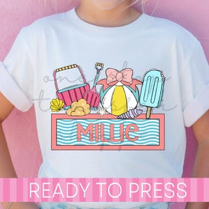 May include: White t-shirt featuring a beach-themed graphic with a pink bucket, shovel, beach ball, popsicle, and the name "Millie" in a pink and blue frame. The words "READY TO PRESS" are printed at the bottom in pink.