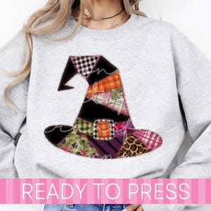 May include: A light gray crewneck sweatshirt featuring a large, central graphic of a witch hat constructed from various patchwork fabric patterns, including plaid, leopard print, floral, and solid colors, all outlined with a dark stitched border.