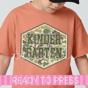 May include: A short-sleeved crew neck t-shirt in a muted orange color. The center of the shirt features a hexagonal graphic with a camouflage pattern in shades of green, brown, and tan. The text "KINDERGARTEN" is printed in dark green block letters.