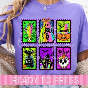 May include: A lavender t-shirt featuring a graphic print of six rectangular postage stamp designs. The colorful illustrations include a hand, a ghost with a cat, a pumpkin, a tombstone with R.I.P. text, a haunted house, and a skull with a candle.