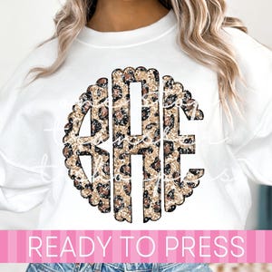 May include: White sweatshirt featuring a large, circular monogram with the letters "GHE" in a leopard print design. The monogram is outlined with a scalloped edge. The text "READY TO PRESS" is displayed in a pink banner across the bottom.