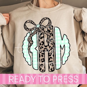 May include: A beige crewneck sweatshirt featuring a central graphic of a monogram with three letters. The design incorporates a leopard print pattern on the letters and a bow, layered over a light green and white gingham circular background.
