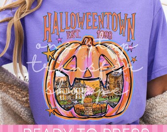 May include: A purple short-sleeve t-shirt featuring a graphic of a large orange jack-o'-lantern with a house scene inside. The text reads HALLOWEENTOWN EST. 1998. The design includes small pink and yellow stars and a watermark overlay.