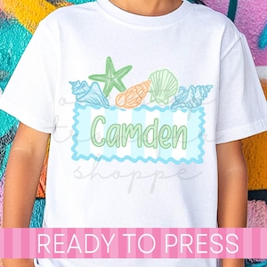 May include: White t-shirt featuring a design with seashells and a starfish in shades of green, blue, and orange. The name "Camden" is displayed in green within a blue striped frame. The text "READY TO PRESS" is printed at the bottom.