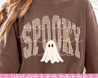 May include: A brown long-sleeve sweatshirt featuring the word SPOOKY in a brown and white gingham pattern. Below the text is a graphic of a white ghost with black eyes. The sweatshirt has a crew neckline and a relaxed fit design.