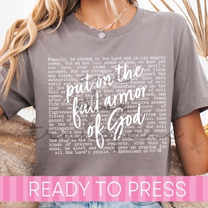 May include: A charcoal gray short-sleeve t-shirt featuring white text that reads "put on the full armor of God" in a large script font, layered over the full text of Ephesians 6:10-18 in a smaller, block-style font across the chest area.