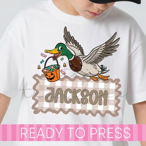 May include: A white t-shirt featuring a graphic of a flying mallard duck holding an orange jack-o'-lantern candy bucket filled with colorful sweets. Below the duck is a beige and white checkered rectangular frame with the name JACKSON.