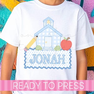 May include: A white t-shirt featuring a graphic of a light blue gingham schoolhouse, a yellow pencil, a red apple, and a blue polka-dot rectangular patch with the name JONAH printed in blue capital letters across the center of the design.
