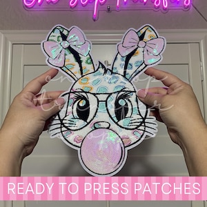 May include: A heat transfer patch featuring a cartoon rabbit with a leopard print pattern, wearing black-rimmed glasses and a light pink bow on each ear, while blowing a large, iridescent pink bubble gum bubble, with a white border outline.