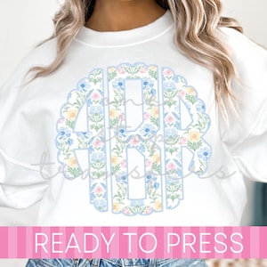 May include: White sweatshirt with a floral monogram design in a scalloped circle. The monogram is filled with blue, pink, and yellow flowers. The text "READY TO PRESS" is displayed on a pink banner.