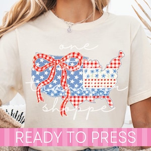 May include: Cream-colored t-shirt featuring a patriotic design. The graphic includes a map of the United States with a red and white gingham bow, stars, and floral patterns. The text "one" and "shoppe" are written in cursive.