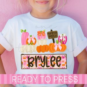 May include: A white t-shirt featuring a graphic of stacked pumpkins in shades of pink, orange, and white, accented with bows. A sign reads PUMPKIN PATCH. Below, a rectangular box with a pink pumpkin pattern displays the name BRYLEE in dark brown text.