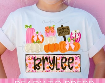 May include: A white t-shirt featuring a graphic of stacked pumpkins in shades of pink, orange, and white, accented with bows. A sign reads PUMPKIN PATCH. Below, a rectangular box with a pink pumpkin pattern displays the name BRYLEE in dark brown text.
