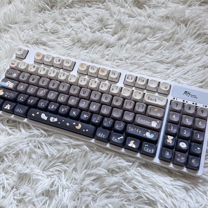 May include: A white mechanical keyboard with a gradient keycap design, transitioning from white to dark gray. The keys feature various icons and illustrations, including moons, stars, and animal paw prints. The keyboard has a numeric keypad on the right side.