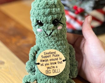 May include: Green crocheted pickle plush toy with a smiling face and black eyes. The toy is holding a wooden sign that reads "Emotional Support Pickle Always around to let you know that you're a BIG DILL!"
