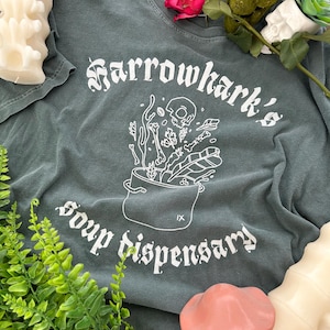 Puede incluir: Una camiseta verde oscuro con un diseño gráfico blanco. El diseño presenta una calavera y huesos en una olla con el texto "Harrowhark's Soup Dispensary".