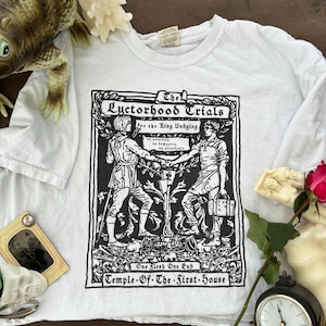 Peut inclure: T-shirt blanc avec une illustration en noir et blanc de deux personnages dans un décor médiéval. Le texte dit "The Luctorhood Trials for the Ring Undying" et "One Flesh One End Temple-Of-The-First-House".