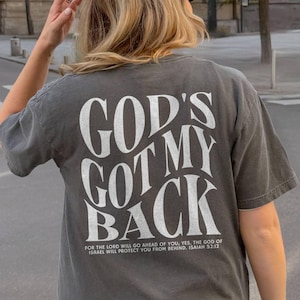 May include: A gray t-shirt with the text "GOD'S GOT MY BACK" in large white letters. Below the text is a smaller line of text that reads "FOR THE LORD WILL GO AHEAD OF YOU, YES, THE GOD OF ISRAEL WILL PROTECT YOU FROM BEHIND. ISAIAH 52:12".