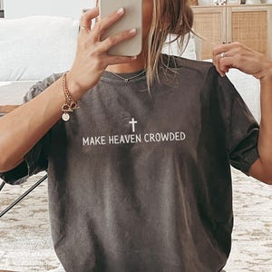 May include: A dark gray t-shirt with the text "MAKE HEAVEN CROWDED" and a cross graphic in white. The shirt is worn with white shorts and black and white striped socks. The person is holding a phone.