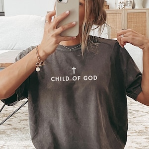 May include: A dark gray t-shirt with the text "CHILD OF GOD" and a cross graphic in white. The shirt is a casual style, suitable for everyday wear. The person is wearing white shorts and white socks with black stripes.